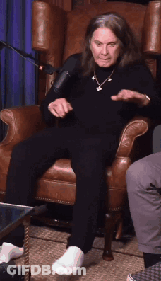 A Man With Long Hair Is Sitting In A Chair With His Feet Crossed GIF
