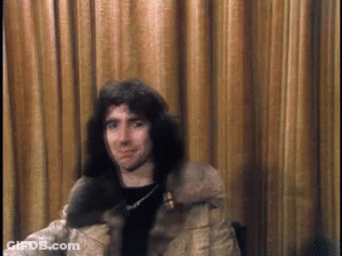 A Man With Long Hair Is Sitting In Front Of A Brown Curtain GIF