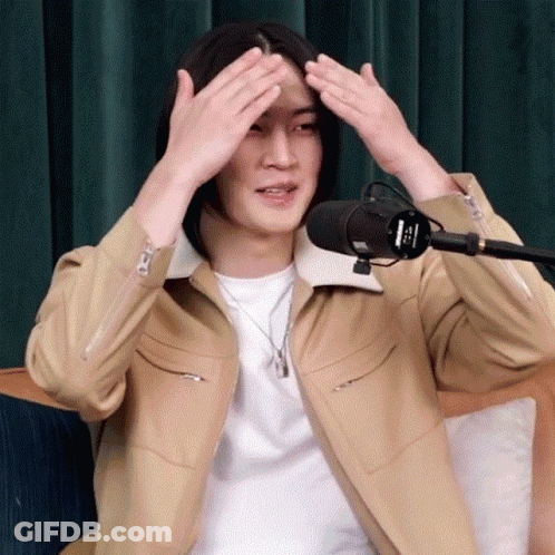 A Man With Long Hair Is Sitting In Front Of A Microphone And Covering His Head With His Hands . GIF