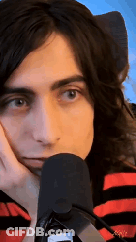 A Man With Long Hair Is Sitting In Front Of A Microphone With His Hand On His Chin . GIF