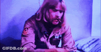 A Man With Long Hair Is Sitting On A Bed Smoking A Cigarette GIF