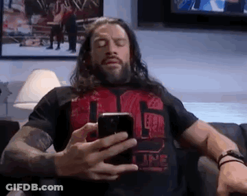 A Man With Long Hair Is Sitting On A Couch Looking At His Phone . GIF