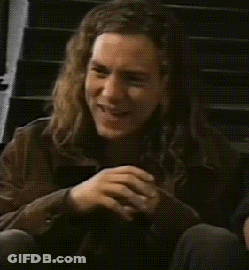 A Man With Long Hair Is Smiling And Sitting On A Set Of Stairs GIF