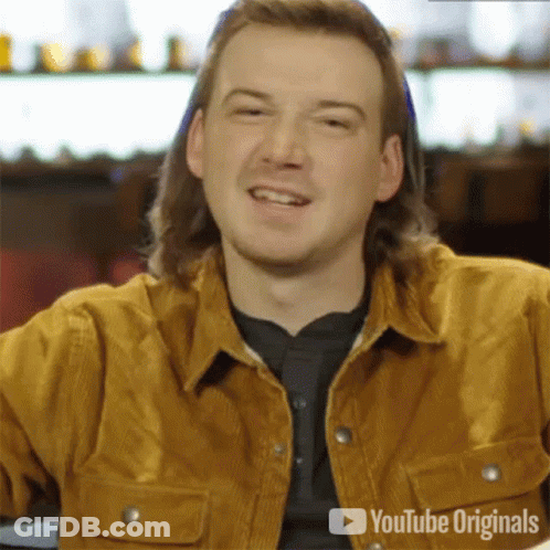 A Man With Long Hair Is Smiling And Wearing A Brown Jacket . GIF