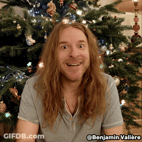 A Man With Long Hair Is Smiling In Front Of A Christmas Tree With The Name Benjamin Valiere On The Bottom GIF