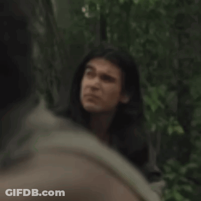 A Man With Long Hair Is Standing In A Forest Looking At Another Man . GIF
