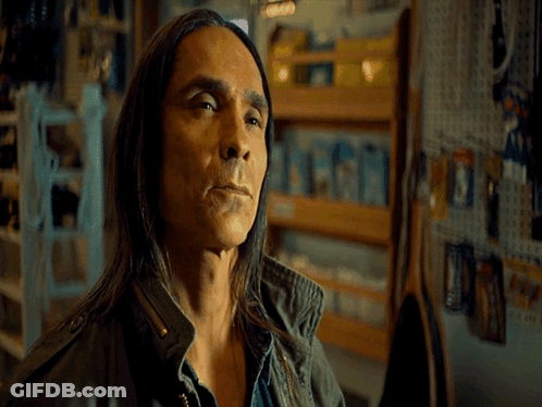 A Man With Long Hair Is Standing In Front Of A Wall That Says Okay GIF