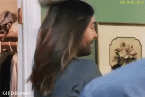 A Man With Long Hair Is Standing In Front Of A Wall With A Picture Of Flowers On It GIF
