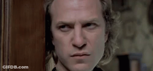 A Man With Long Hair Is Standing In Front Of A Wooden Door Looking At The Camera . GIF