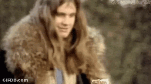 A Man With Long Hair Is Wearing A Fur Coat And Holding A Glass Of Water . GIF