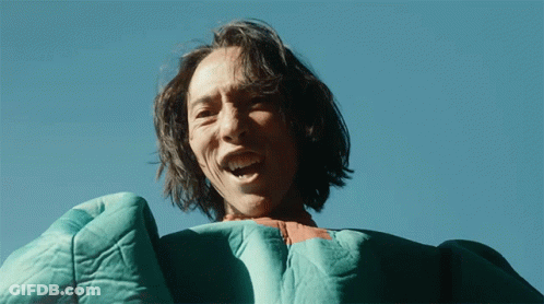 A Man With Long Hair Is Wearing A Green Blanket And Smiling GIF
