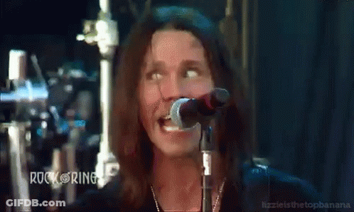 A Man With Long Hair Singing Into A Microphone With Rock Ring Written On The Bottom Left GIF