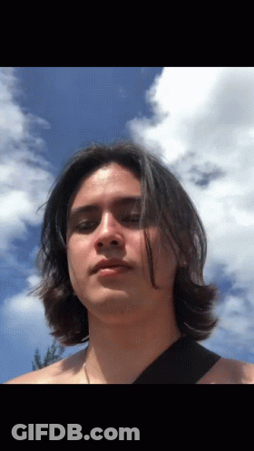 A Man With Long Hair Stands In Front Of A Blue Sky With Clouds GIF