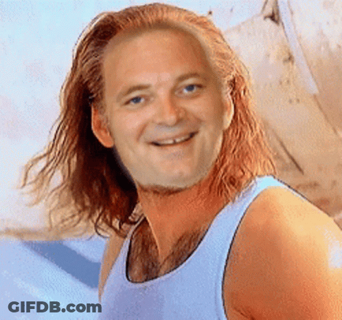 A Man With Long Red Hair Wearing A White Tank Top Smiles For The Camera GIF
