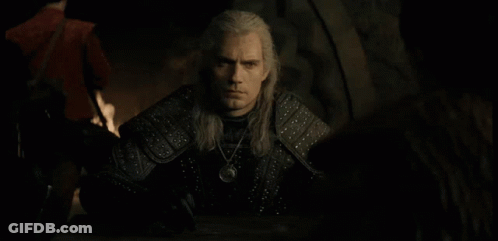 A Man With Long White Hair Is Sitting At A Table With A Necklace Around His Neck GIF
