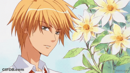 A Man With Orange Hair Is Standing In Front Of A Bush Of Flowers GIF