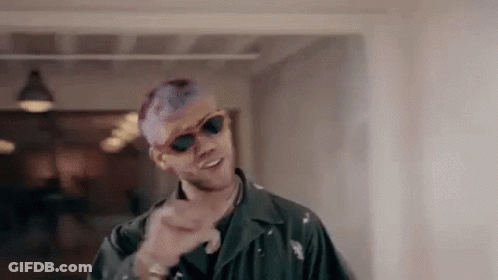 A Man With Purple Hair And Sunglasses Is Standing In A Hallway And Pointing At The Camera . GIF