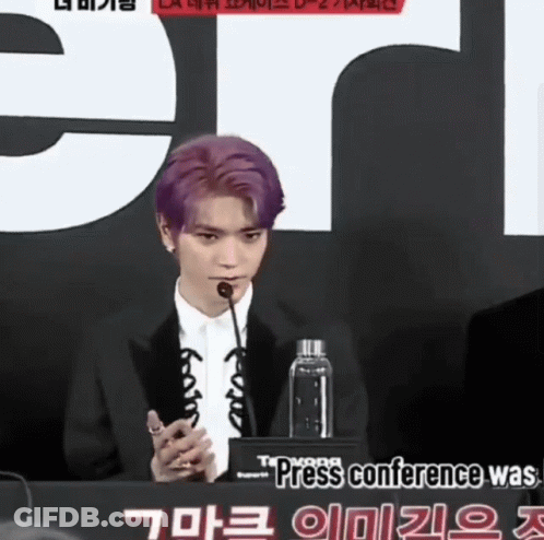 A Man With Purple Hair Speaking Into A Microphone In Front Of A Sign That Says Press Conference GIF