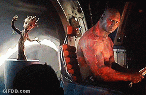 A Man With Red Paint On His Body Is Sitting In A Chair Next To A Statue Of Groot . GIF