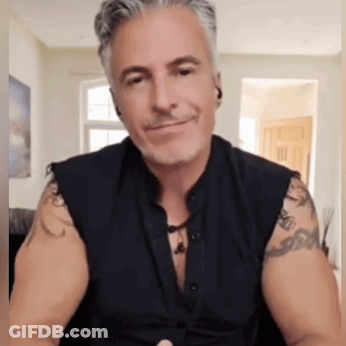 A Man With Tattoos On His Arms Is Wearing A Black Vest GIF