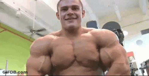 A Man With Very Large Muscles Is Standing In A Gym And Smiling At The Camera . GIF