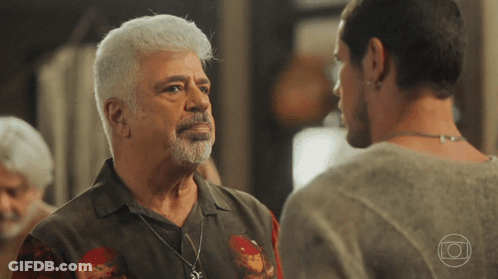 A Man With White Hair And A Beard Stands Next To Another Man With A Necklace Around His Neck GIF
