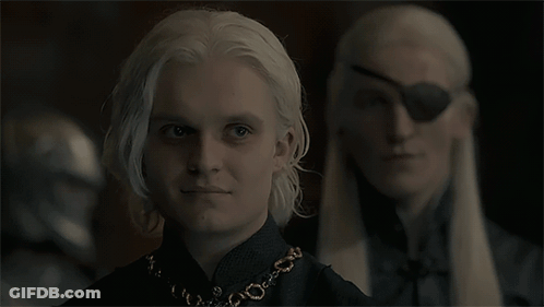 A Man With White Hair And A Necklace Is Standing Next To Another Man With A Blindfold On His Eye GIF