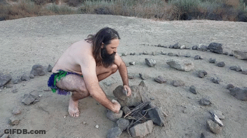 A Man Without A Shirt Is Kneeling In The Dirt Near A Rock Labyrinth GIF