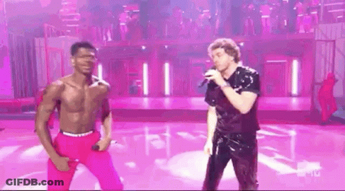 A Man Without A Shirt Is Singing Into A Microphone While Another Man Stands Next To Him On A Stage . GIF