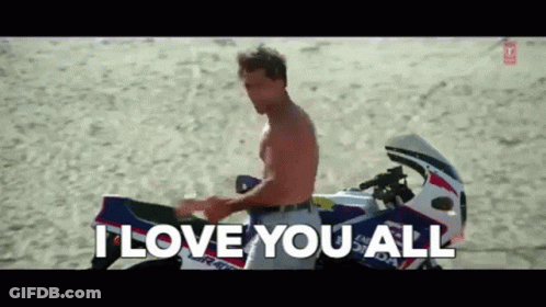 A Man Without A Shirt Is Sitting On A Motorcycle On The Beach And Saying I Love You All . GIF