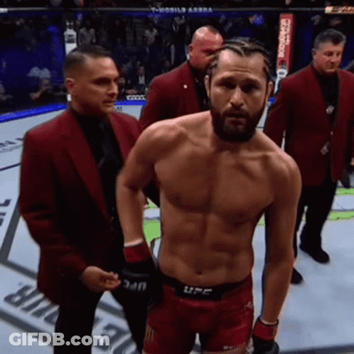 A Man Without A Shirt Is Standing In A Boxing Ring Surrounded By Men In Red Suits . GIF
