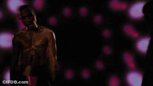 A Man Without A Shirt Is Standing In A Dark Room With Purple Lights Behind Him GIF