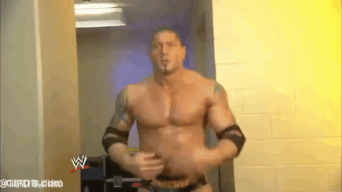A Man Without A Shirt Is Standing In A Hallway With A Wrestling Logo On The Wall Behind Him . GIF