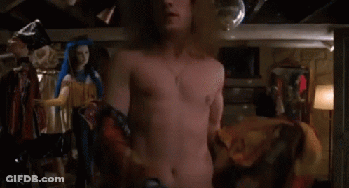 A Man Without A Shirt Is Standing In A Room With A Woman In A Costume Behind Him . GIF