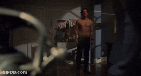 A Man Without A Shirt Is Standing In A Room With Posters On The Wall GIF