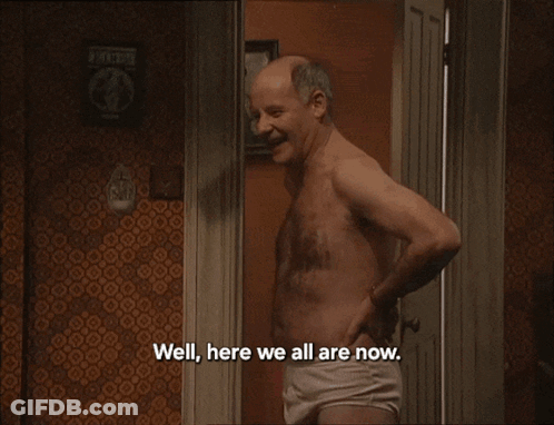 A Man Without A Shirt Is Standing In Front Of A Bed With The Words Well Here We All Are Now All The Lads GIF