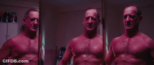 A Man Without A Shirt Is Standing In Front Of A Mirror And Looking At Himself GIF
