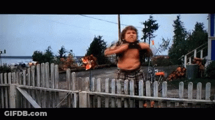 A Man Without A Shirt Is Standing In Front Of A Wooden Fence Holding A Windmill . GIF
