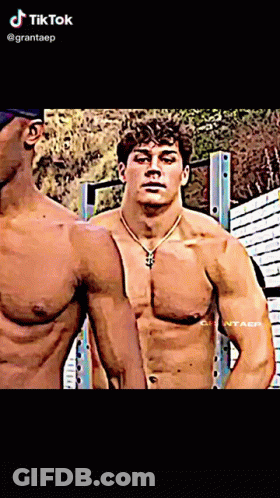 A Man Without A Shirt Is Standing Next To Another Shirtless Man With A Necklace Around His Neck .. GIF