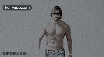 A Man Without A Shirt Is Standing On A Beach Wearing Sunglasses . GIF