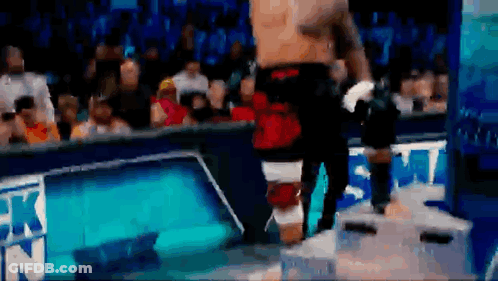 A Man Without A Shirt Is Standing On A Box In Front Of A Crowd At A Wrestling Match . GIF