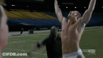 A Man Without A Shirt Is Standing On A Football Field While Another Man Looks On . GIF