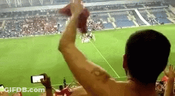 A Man Without A Shirt Is Taking A Picture Of A Soccer Game While A Crowd Watches . GIF