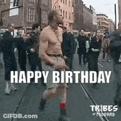 A Man Without A Shirt Is Walking Down A Street In Front Of A Crowd Of People . GIF