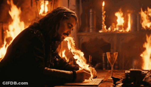 A Man Writes On A Piece Of Paper In Front Of A Fireplace With Flames Coming Out Of His Mouth GIF