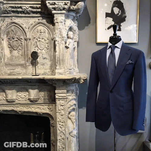 A Mannequin Wearing A Blue Suit And Tie Stands In Front Of A Stone Fireplace GIF