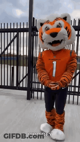 A Mascot Dressed As A Tiger Is Standing In Front Of A Fence With His Arms Crossed . GIF