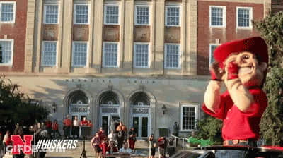 A Mascot For The Nebraska Huskers Stands In Front Of A Large Building GIF