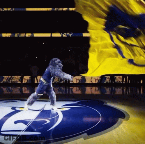 A Mascot Is Dancing On A Basketball Court With A Yellow Flag GIF
