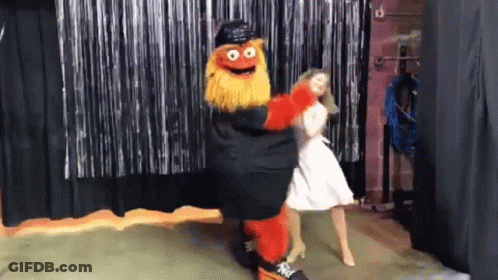 A Mascot Is Dancing With A Woman In A White Dress In Front Of A Black Curtain GIF